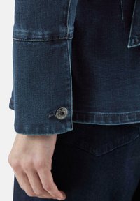 Donkerblauwe spijkerjackmouw met een zichtbare zilveren drukknop. Versleten textuur en stiksels langs de rand van de manchet. Hand deels zichtbaar.