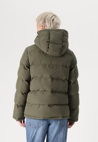Olijfgroene pufferjack met een capuchon, voorzien van baffle-stiksel voor isolatie en een relaxed fit. Gecombineerd met lichtblauwe jeans.