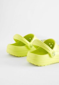 Félpár élénk sárga, slip-on klumpa állítható hátrésszel és szellőző lyukakkal, sima fehér háttér előtt.