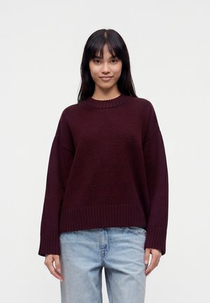 Kvinde med langt sort hår iført en mørkelilla sweater og lyseblå jeans, stående mod en ensartet lys baggrund.