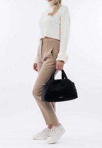 Sac en cuir noir à texture pierre, avec de courtes poignées et une forme incurvée, tenu dans une main à côté d'un pantalon taille haute beige et de baskets blanches.