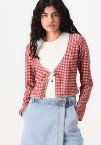 Veste cropped en vichy rouge et blanc avec des manches longues, une dentelle le long de l'encolure et un détail à nouer à l'avant. Associée à une jupe en denim clair.