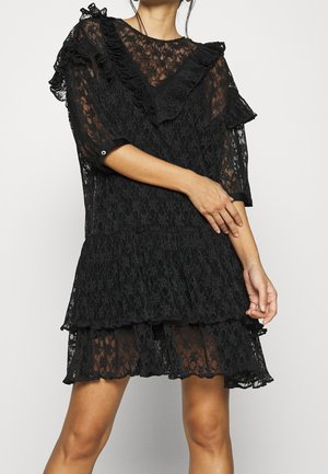 Femme portant une robe noire en dentelle avec des volants en couches et des manches transparentes, posant avec un bras croisé sur son corps.