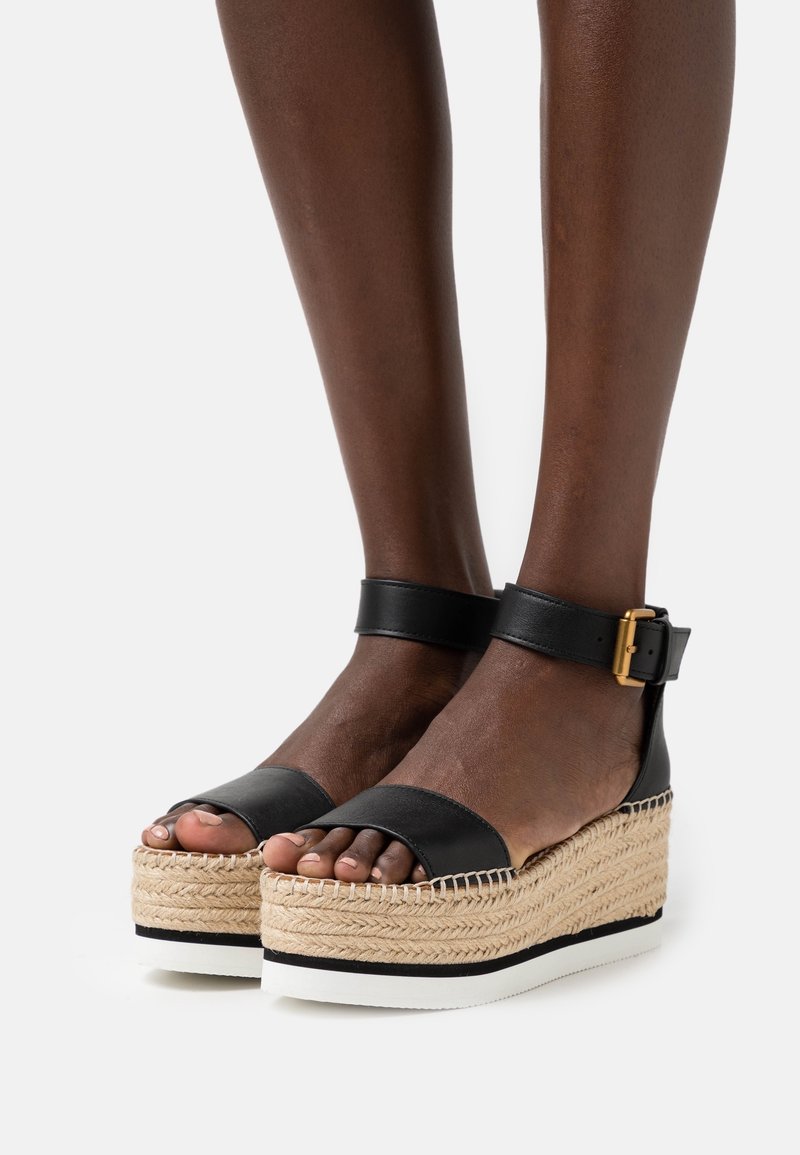 Sandales à plateforme en cuir noir avec une tige en jute tissée, bout ouvert, bride de cheville et détail de boucle en métal doré. Accents de semelle blanche et noire.