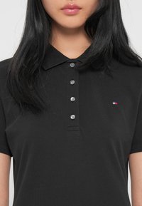 Sort polo-shirt lavet af tekstureret stof, med en skjorteskrave, seks grå knapper og et lille logo i rød, hvid og blå.