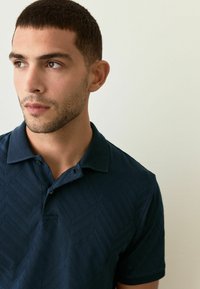 Cămașă polo bleu închis, cu un model geometric texturat, mâneci scurte și guler cu nasturi. Materialul pare neted și respirabil.