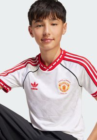 Bijela Manchester United majica s crvenim akcentima, s geometrijskim uzorkom, crvenim ovratnikom i tri crvene pruge na ramenima.