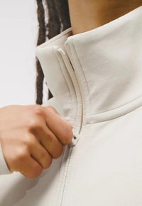 Svetlý zipsový kabát s vysokým golierom, s viditeľným zipsom a mäkkou, hladkou textúrou. Ruka drží za ťaháčik zipsu.