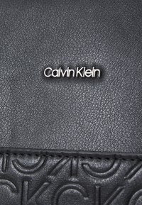 Černá kožená taška s vyraženými logovými akcenty, texturovaným povrchem, hladkým šitím a výrazným kovovým emblémem Calvin Klein.