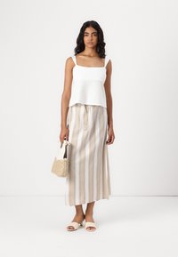 Top blanc sans manches, court, associé à une longue jupe rayée beige et blanche. Accessoirisé avec un petit sac tissé et des sandales à enfiler.