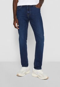 Vaqueros de mezclilla azul oscuro con un corte ajustado, con costuras naranja y un diseño clásico de cinco bolsillos, combinados con zapatillas blancas.