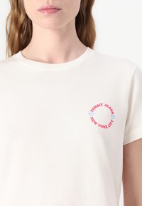Kremo spalvos trumpas rankovėmis marškinėliai su apvaliu piešiniu. Pateiktas tekstas: "TOMMY JEANS" raudona ir "NEW YORK CITY" mėlyna spalvomis su širdies akcentais.