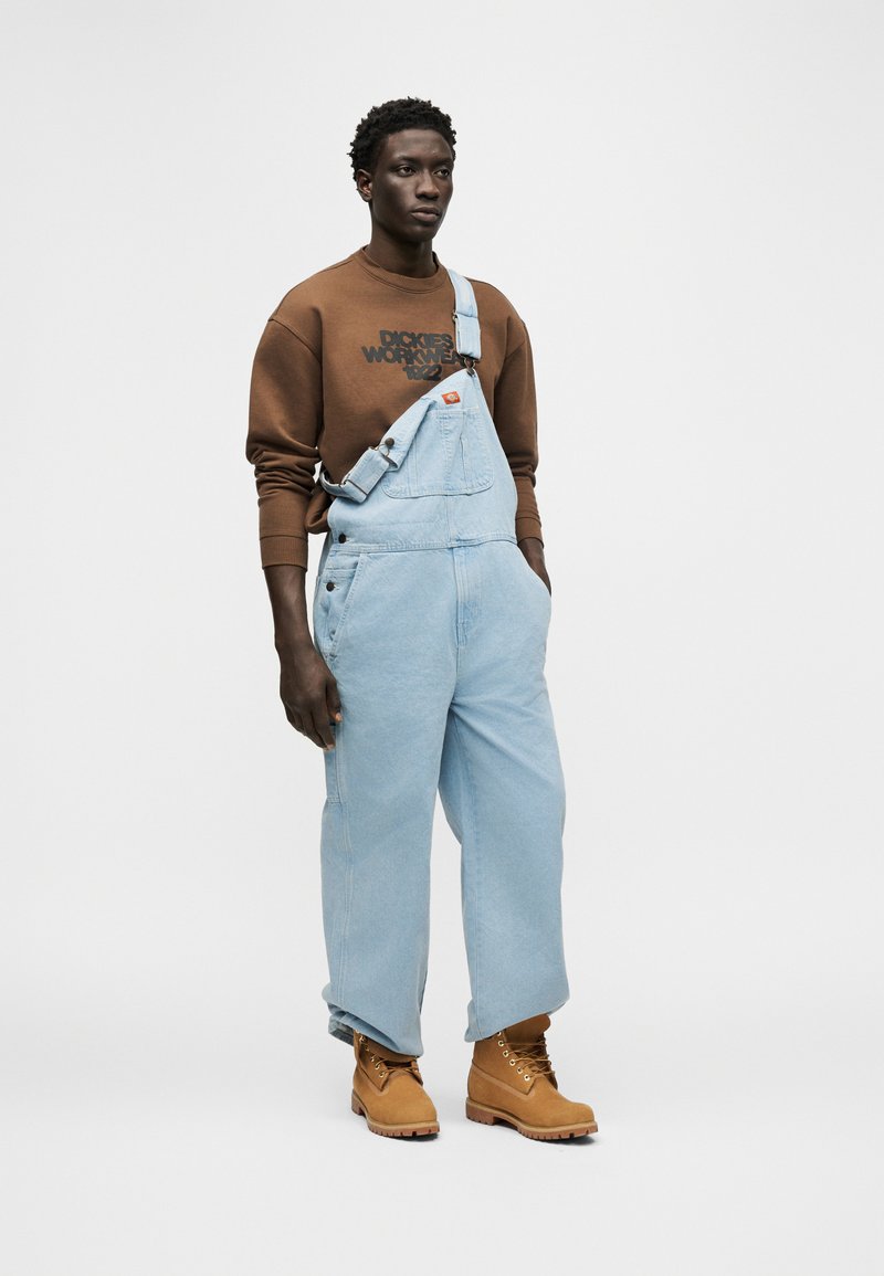 Junger Mann, der hellblaue Jeans-Latzhosen mit einem befestigten Träger, ein braunes Sweatshirt und hellbraune Arbeitsstiefel trägt und vor einem einfarbigen Hintergrund steht.