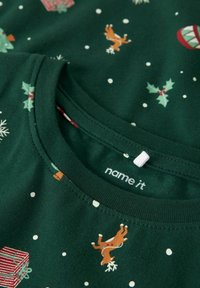 Groene katoenen T-shirt met een feestafbeelding van rendieren, cadeaus en hulst, met witte stippen en een geribbelde halslijn. Merkenlabel zichtbaar.