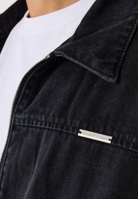 Chaqueta de mezclilla negra con collar, cremallera plateada y una pequeña etiqueta metálica grabada "PROJECT X PARIS" en la zona del pecho. Tela texturizada.