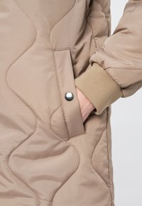 Izšūts bēšas krāsas džemperis ar ribotām aprocēm, aprīkots ar sānu kabatu ar sudraba pogu un gludu, nedaudz spīdīgu auduma tekstūru.