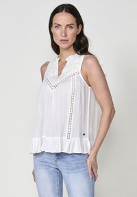 Blusa senza maniche bianca con scollo alto, dettagli ricamati e accentuazioni in pizzo. Caratterizzata da un orlo arricciato e un tessuto leggero e testurizzato.