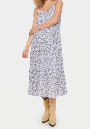 Kvinde iført en ærmeløs, hvid blomster-middagkjole sammen med lysebrune ruskind-cowboystøvler, stående mod en ensfarvet hvid baggrund.