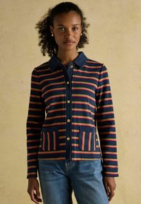 Prúžkovaná bunda v námorníckej modrej, červenej a krémovej farbe, s golierom, zlatými gombíkmi a dvoma prednými vreckami. Mäkko tkanina s jemne textúrovaným povrchom.