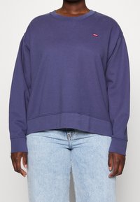 Persona con una felpa viola di Levi's a maniche lunghe abbinata a jeans chiari a vita alta, in piedi su uno sfondo neutro.