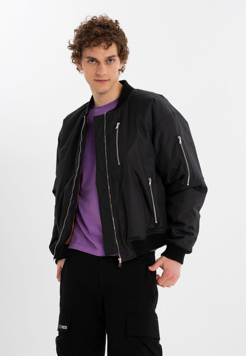 Black bomber jacket with zippered pockets, ribbed cuffs, and a quilted inner lining, worn over a purple shirt and black pants.