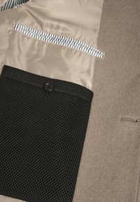 Interior de un abrigo beige que muestra un bolsillo de malla negra con botón, un bolsillo interior a rayas y un exterior de tela de lana con ojal.