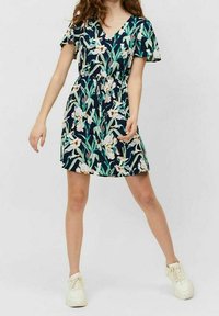 Blumenkleid mit V-Ausschnitt, kurzen Ärmeln, tailliertem Schnitt und dunkelblauem Hintergrund. Es zeigt grüne Blätter und cremefarbene Blumen; getragen mit hellen Sneakers.