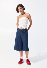 Denim-Culottes mit weiter Beinform, tiefblauer Farbe und kontrastierenden Nähten. Kombiniert mit einem figurbetonten weißen trägerlosen Oberteil und roten Sneakern.