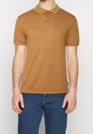 Mann trägt ein braunes kurzärmeliges Poloshirt mit drei Knöpfen, kombiniert mit blauen Jeans, steht vor einem schlichten hellen Hintergrund.