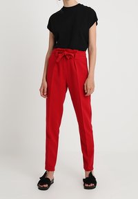 Red high-waisted trousers with a fabric tie belt, tailored fit, and smooth texture, paired with a black short-sleeve shirt and black sandals.