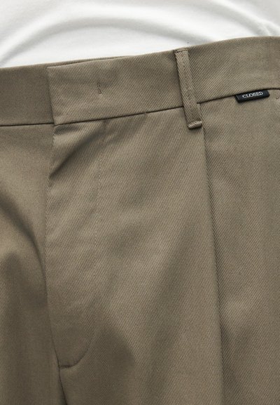 Gros plan sur un pantalon beige avec passants de ceinture, poche latérale et petite étiquette de la marque "CLOSED" près de la ceinture, porté avec un haut blanc.