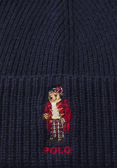 Marineblaue Strickmütze mit geripptem Design und einem gestickten Bären in einem roten Mantel mit Karohosen sowie einem roten Akzent darunter.