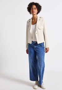 Cremfarbener Blazer mit zwei vorderen Taschen, kragenloser Gestaltung und Knopfverschluss, kombiniert mit lockeren blauen Jeans und weißen Sneakers.