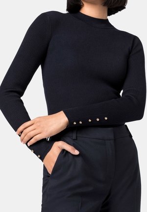 Femme portant un haut noir moulant en côtes à manches longues avec des boutons dorés aux poignets et un pantalon noir taille haute, main dans la poche.