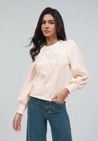 Lys rosa hettegenser med lange pufferm, pyntet med en sløyfe designet med små perler og en cropped kant.