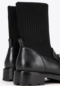 Botas de tobillo de cuero negro con un upper de punto negro acanalado, que presentan una punta redondeada, suela gruesa y una tirador adicional para facilitar el calzado.