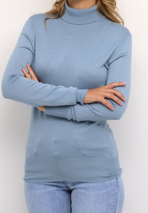 Donna che indossa un maglione dolcevita azzurro chiaro e jeans a lavaggio chiaro, con le braccia incrociate sul petto contro uno sfondo chiaro e uniforme.