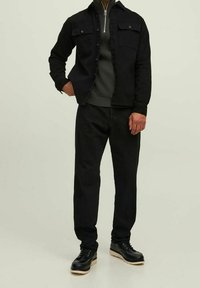 Chaqueta de denim negra con bolsillos de solapa, superpuesta sobre un suéter de canalé verde oscuro, combinada con pantalones negros y zapatos negros con suelas beige.