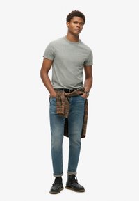 Grijze t-shirt met subtiele textuur, blauwe jeans en zwarte laarzen. Een geruite flanel die om de taille is geknoopt voegt kleur en patrooncontrast toe.