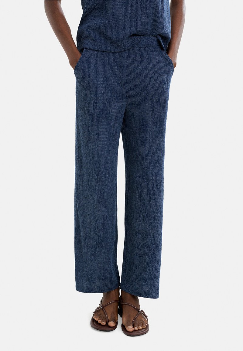 Granatowe spodnie z szerokimi nogawkami wykonane z teksturowanej tkaniny, z elastycznym pasem i bocznymi kieszeniami. Połączone z brązowymi klapkami.