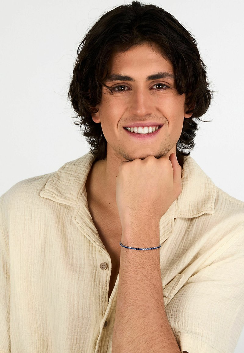 Camicia texturizzata beige chiaro, indossata con la mano appoggiata sotto il mento. Un braccialetto blu con pietre alternate è indossato al polso.