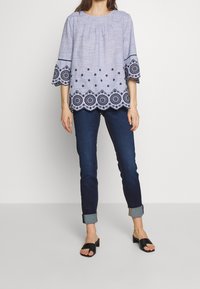 Ljust blå broderad blus med trekvarts bellärmar och vågig nederkant, kombinerad med mörka jeans och svarta slide-sandaler.