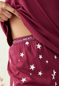 Set de pijama burgund roșu cu un model de stele albe; talie elastică vizibilă cu textul "NEXT iubește"; material moale și ușor.