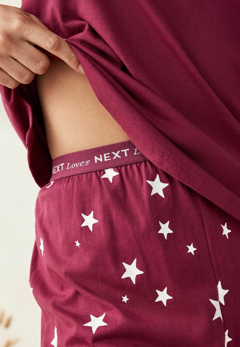 Set de pijama burgund roșu cu un model de stele albe; talie elastică vizibilă cu textul "NEXT iubește"; material moale și ușor.