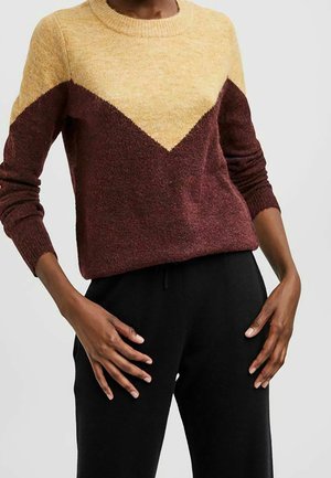 Modelo que lleva un suéter de lana de manga larga con un patrón de chevron en amarillo y burdeos, combinado con pantalones negros.