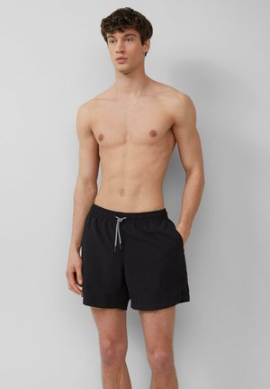 Junger Mann, der shirtlos vor einem schlichten Hintergrund steht, in schwarzen Badeshorts mit einem Frontzugband und Seitentaschen.