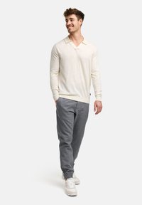 Jasno beżowy longsleeve polo z dekoltem w serek i ściągaczami na rękawach, w połączeniu z szarymi tapered spodniami i białymi trampkami. Gładka faktura, swobodny wygląd.