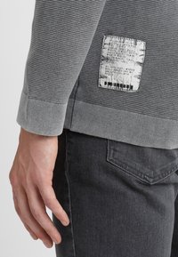 Grauer strukturierter Pullover mit gerippten Bündchen, ausgestattet mit einem rechteckigen Stoffetikett auf der Rückseite, das Pflegehinweise und ein Musterdetail zeigt.