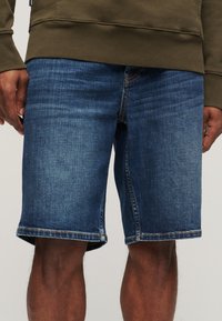 Hombre que lleva pantalones cortos de mezclilla azul de longitud media y sudadera verde oliva, con las manos relajadas a los lados frente a un fondo neutro.
