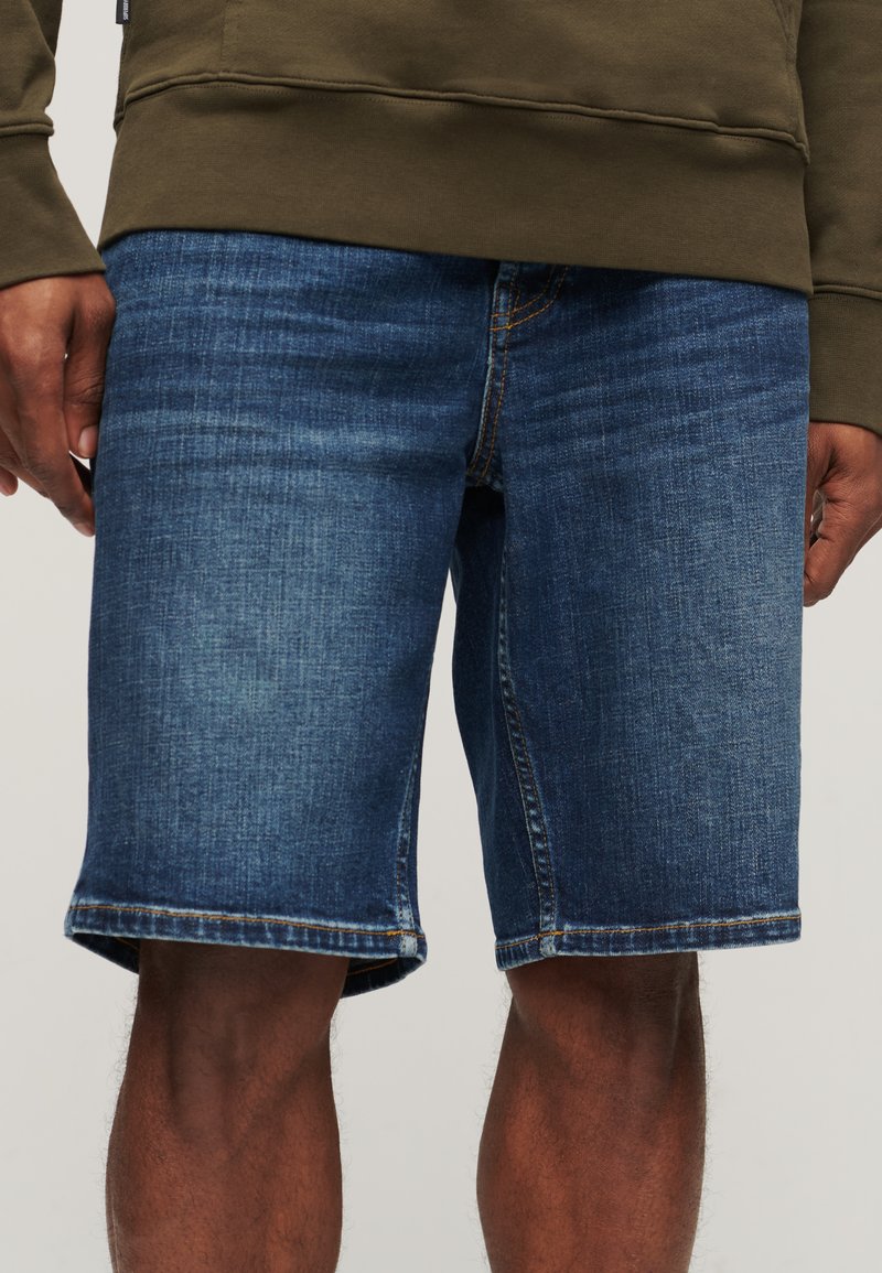 Hombre que lleva pantalones cortos de mezclilla azul de longitud media y sudadera verde oliva, con las manos relajadas a los lados frente a un fondo neutro.